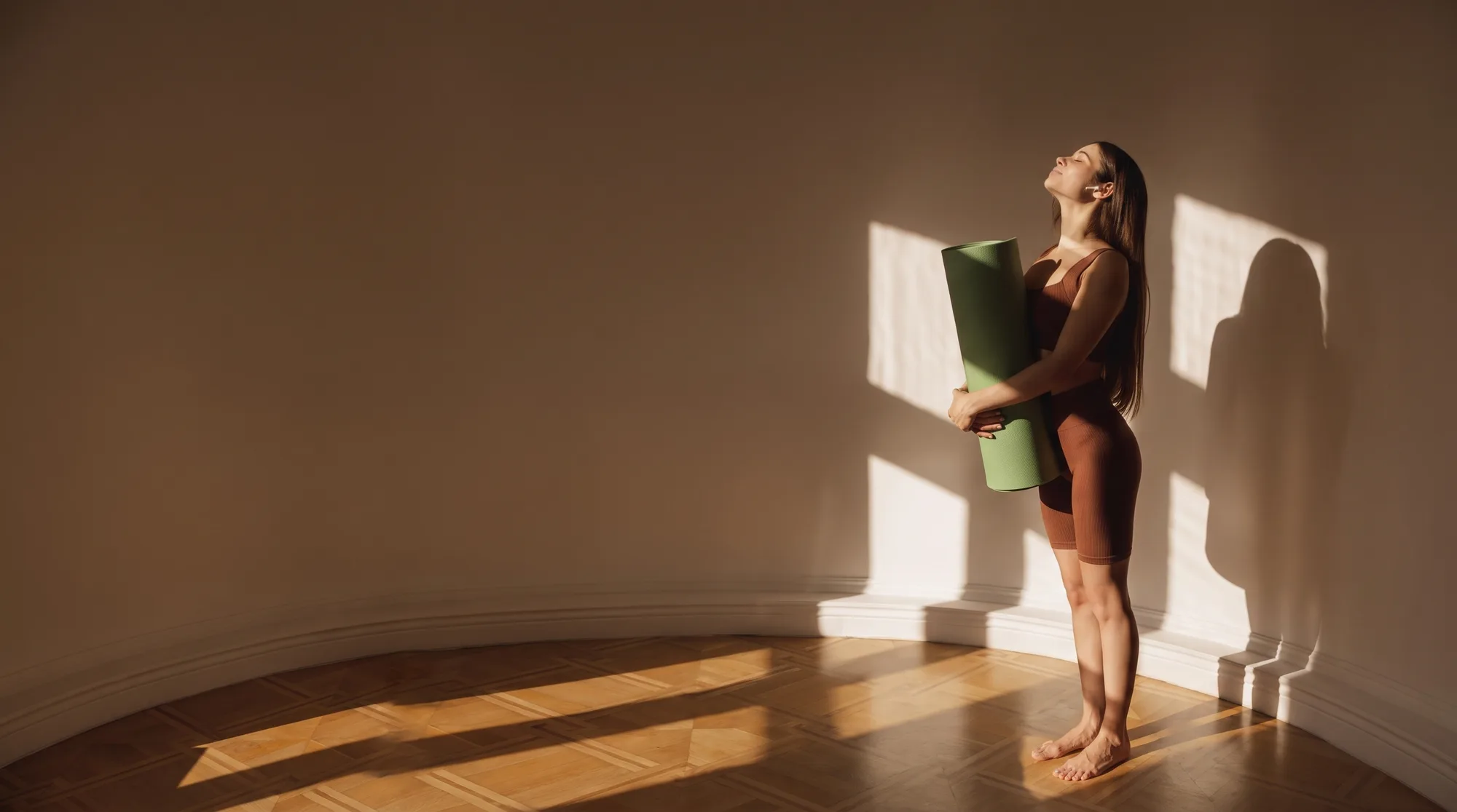 A woman standing barefoot in a sunlit room, holding a rolled yoga mat